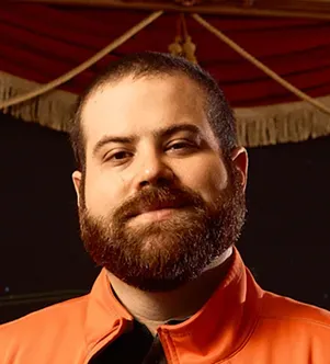 A portrait of a man with a dark beard wearing an orange jacket, standing inside the historic Fulton Theatre in Pennsylvania, featured in a case study about using AI in theatre management.