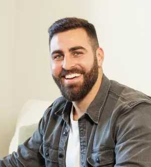 A smiling man with a well-groomed beard and short dark hair, wearing a white t-shirt and an open grey denim button-down shirt, sitting indoors.
