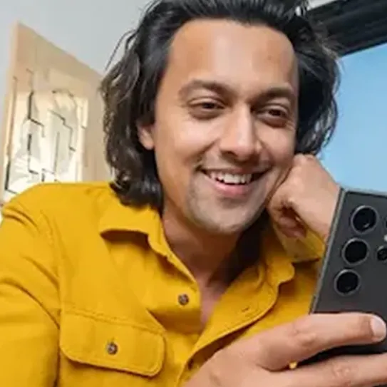 A man with dark, wavy hair and a yellow shirt smiles while using a smartphone. The background is blurred, suggesting a home or casual office setting.