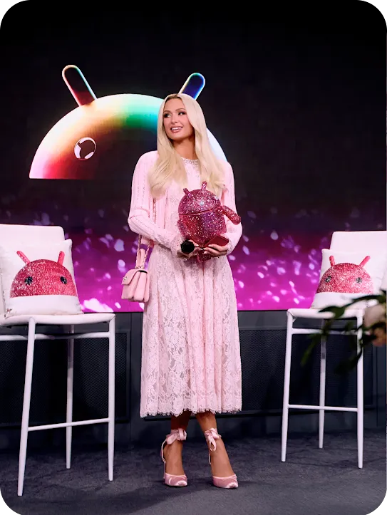Paris Hilton standing on stage in a pink lace dress holding a bedazzled pink Android mascot, with a colorful Android logo illuminated behind her.