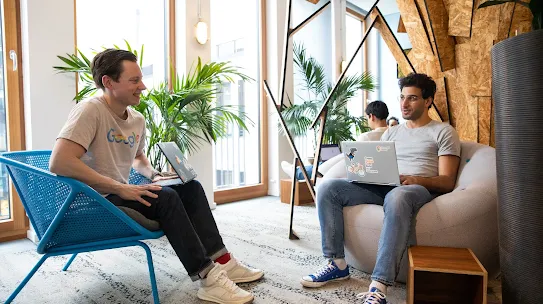 Two colleagues are seated in a bright, modern office lounge, having a casual discussion. On the left, a man wearing a grey Google-branded t-shirt, dark jeans, and white sneakers sits in a bright blue, perforated mesh armchair. On the right, a man with glasses and a grey t-shirt sits in a large, round grey beanbag-style chair, with a laptop open on his lap. Both men have laptops decorated with various colorful stickers. Large windows in the background let in plenty of natural light, and the space is decorated with potted palm plants and a unique architectural wall feature made of textured wood panels.