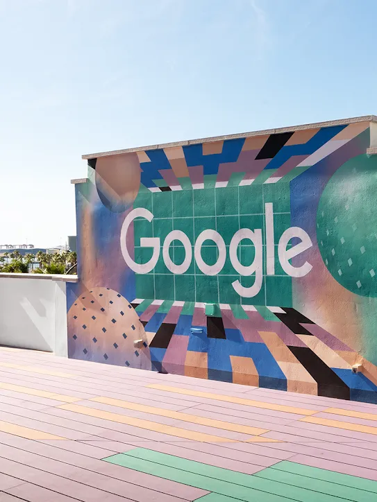 Um terraço com o logotipo do Google em uma parede. Há palmeiras e barcos ao longe.
