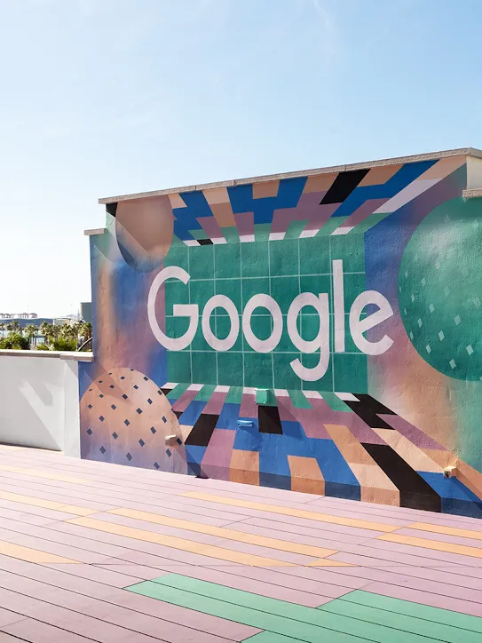 Dach budynku z logo Google na ścianie. W tle widać palmy i statki.