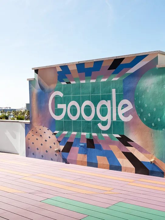 على سطح مبنى، جدارٌ يزيّنه شعار Google، في حين تتناثر أشجار النخيل والقوارب في الأفق