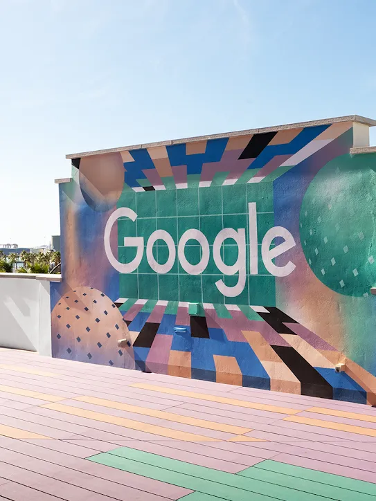 Duvarında Google logosu bulunan bir teras. Uzakta palmiye ağaçları ve tekneler görülüyor.