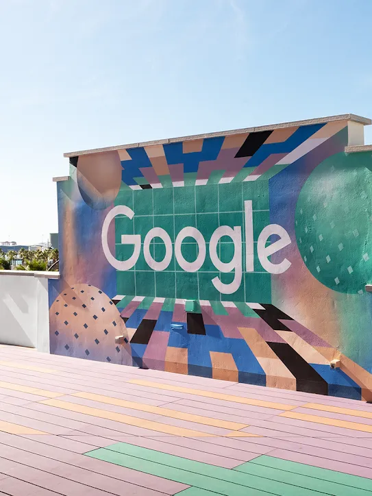 屋顶有一面墙，墙上印有 Google 徽标。远处是棕榈树和船只。