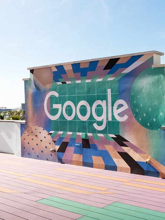 Un toit avec le logo Google sur un mur. On voit des palmiers et des bateaux au loin.