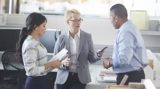 Three professionals standing in a modern office environment engaged in a discussion. A woman in the center, wearing glasses and a grey blazer, holds a smartphone while conversing with a woman on her left holding a folder and a man on her right.