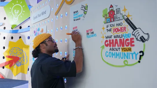 A man wearing a yellow beanie, glasses, and a black shirt writes on a large white interactive wall with an orange marker. On the right side of the wall, colorful illustrated text reads: "IF YOU HAD THE POWER WHAT WOULD YOU CHANGE ABOUT YOUR COMMUNITY?" Next to him, other written responses on the board include "BETTER MONEY MANAGEMENT FOR SCHOOLS" and "BETTER PAY FOR TEACHERS." The background features a brightly colored, interactive exhibit space with a sign that says "Speak Your Mind."