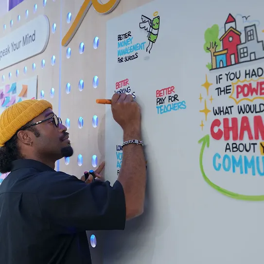 A man wearing a yellow beanie, glasses, and a black shirt writes on a large white interactive wall with an orange marker. On the right side of the wall, colorful illustrated text reads: "IF YOU HAD THE POWER WHAT WOULD YOU CHANGE ABOUT YOUR COMMUNITY?" Next to him, other written responses on the board include "BETTER MONEY MANAGEMENT FOR SCHOOLS" and "BETTER PAY FOR TEACHERS." The background features a brightly colored, interactive exhibit space with a sign that says "Speak Your Mind."