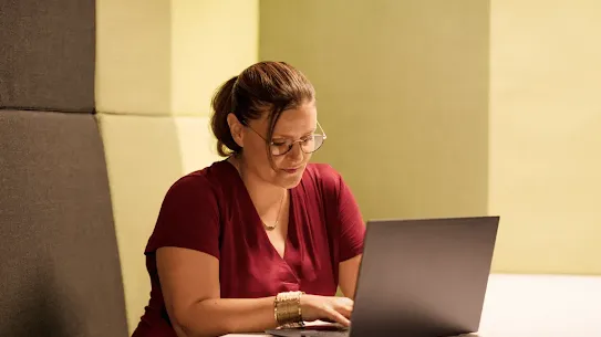 A professional woman with glasses and a red shirt sits in a modern, recessed acoustic work booth, focused on her laptop. The warm, yellow-toned lighting and quiet setting symbolize the focused work required for digital security and transforming cybersecurity in the EU.