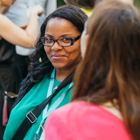 A dark woman with long hair and wearing spectacles smiling at other woman in crowd.
