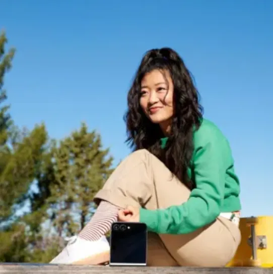 The Samsung Galaxy Z Flip6 is unfolded halfway so that it’s propped upright. It’s facing a person happily sitting outside and carrying out a conversation on a sunny day.