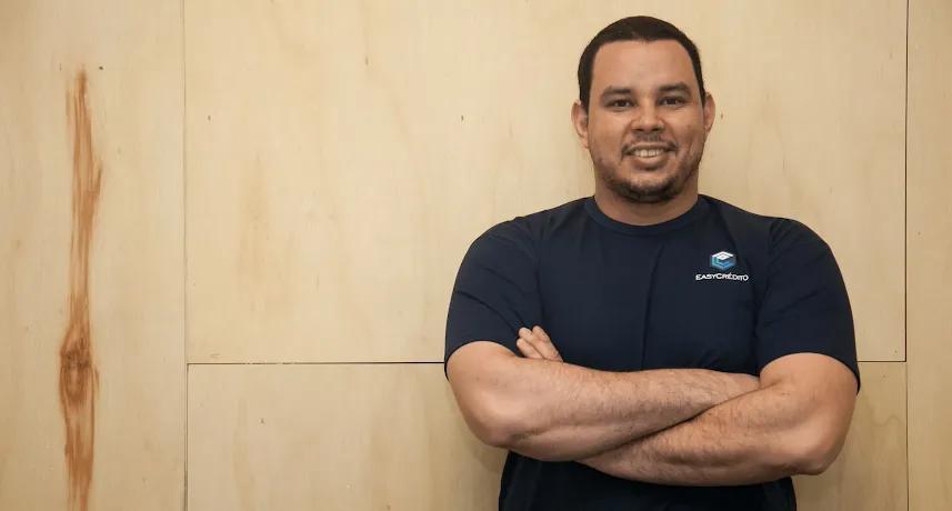 Homem em pé, de braços cruzados, em frente a fundo de madeira.