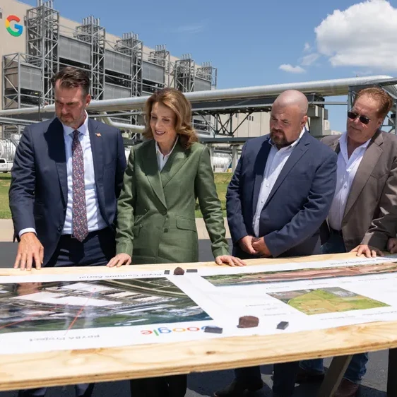 A group of six people, five men and one woman, standing around printed plans of a data center site on a wood table