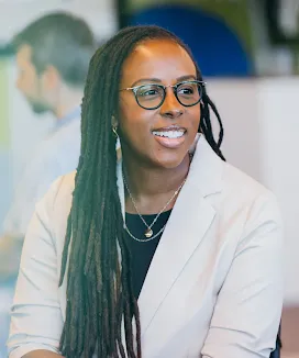 A smiling professional wearing glasses and a white blazer is engaged in conversation in a bright office setting.