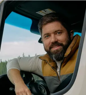 A smiling man with a beard sits in the driver's seat of a Mercedes-Benz sprinter van, leaning his arm on the steering wheel with evergreen trees visible in the background.