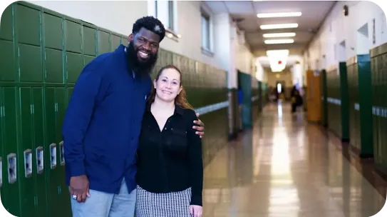 A man with a full dark beard and a navy sweater stands with his arm around a smiling woman in a long-sleeved black shirt, posing together in a long school corridor lined with green lockers.