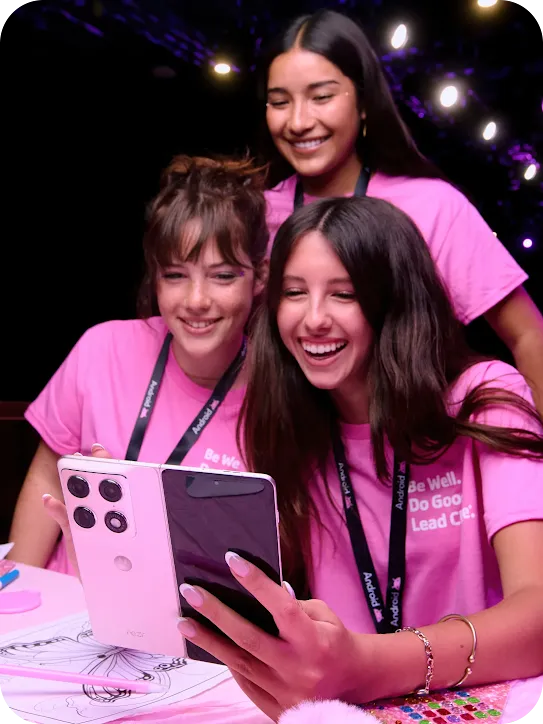 Three smiling girls in pink shirts sitting at a pink craft table, looking joyfully at a white folding smartphone held by one of them.