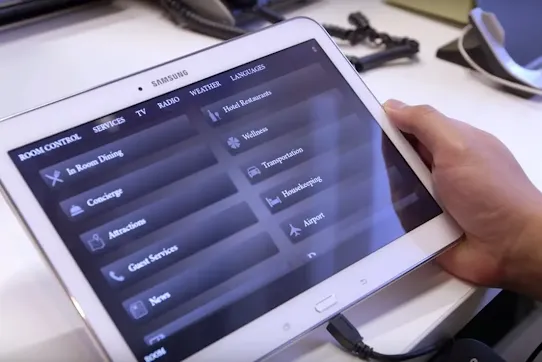 A person holding a Samsung tablet that displays a hotel service menu, including options for In-Room Dining, Wellness and Transportation.