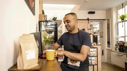 A coffee shop owner in an apron, works at his laptop while sipping his morning coffee from a bright yellow cup.