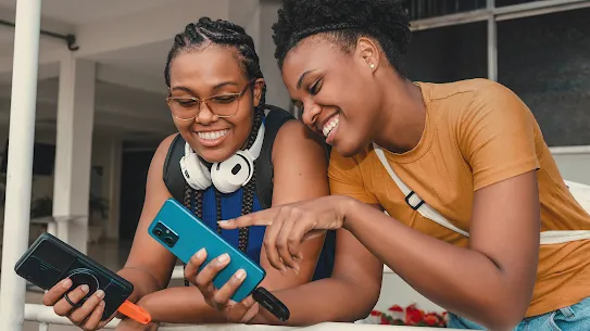 Two smiling young women outdoors looking at and pointing to a smartphone screen together.
