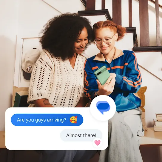 Two people are sitting down looking at one of their text messages. The text message exchange overlays the picture. There's a blue text bubble that reads &quot;are you guys arriving?&quot; next to a smiling emoji with hearts. Underneath is is a gray text bubble that reads &quot;Almost there!&quot; with a pink heart reaction. A Google Messages icon hovers on the upper left corner of the text box.