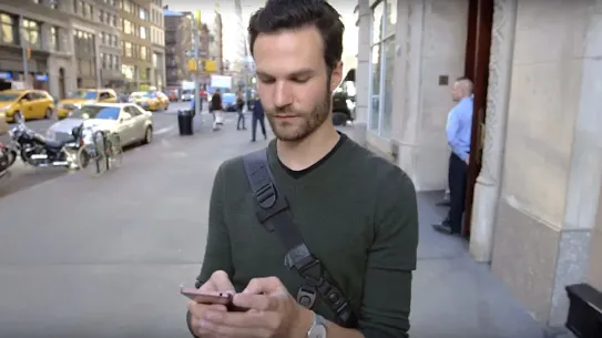 A man with a crossbody bag using smartphone on a city sidewalk.