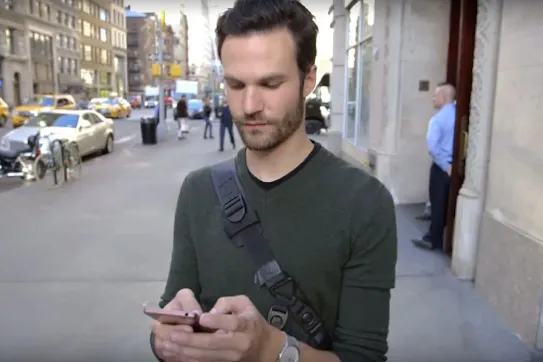 A man with a crossbody bag using smartphone on a city sidewalk.