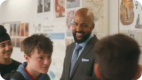 A smiling Black male teacher in a grey suit and blue tie stands in a classroom, looking warmly at a group of diverse students. Educational posters about Egypt and history are visible on the wall in the background.