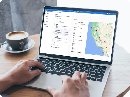 A close-up, over-the-shoulder shot of hands typing on a silver laptop keyboard. The laptop rests on a wooden table next to a cup of coffee in a dark saucer. The screen displays the Google Cloud Platform console, showing the Google Maps Platform "Quick Builder" tool used to create a business locator map. The preview panel on the right displays a map of California marked with several red location pins.
