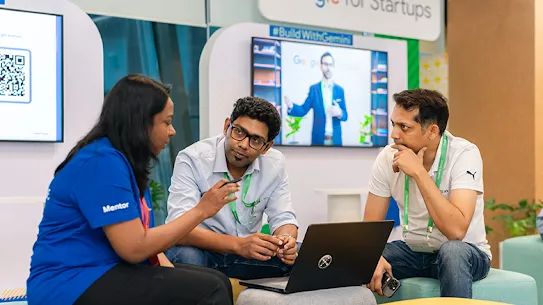 Three professionals sitting on colorful ottomans collaborate around a laptop in a "Google for Startups" branded event space.