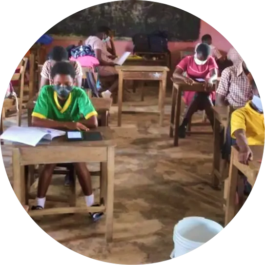 Students wearing face masks study at individual wooden desks inside a classroom with a chalkboard background, cropped into a circular frame.