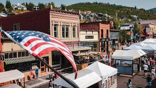 An elevated view of a bustling historic American Main Street during a bright, sunny day. An American flag waves in the foreground, overlooking a street lined with red brick buildings, local shops, and outdoor market tents where people are walking and browsing. Green hills with residential houses are visible in the background.