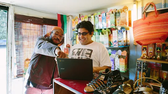 A man points to a computer screen. Another man smiles looking at the screen.
