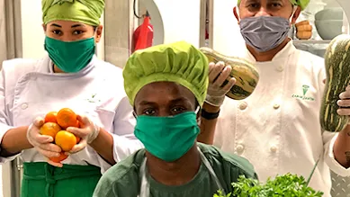 Three chefs in masks and white robes are holding up fresh produce for the camera.
