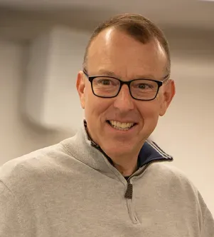 A professional portrait of Dan Kelley, a Rhode Island educator, smiling with glasses and wearing a gray quarter-zip sweater, featured in a case study about using Google AI to reduce administrative paperwork.