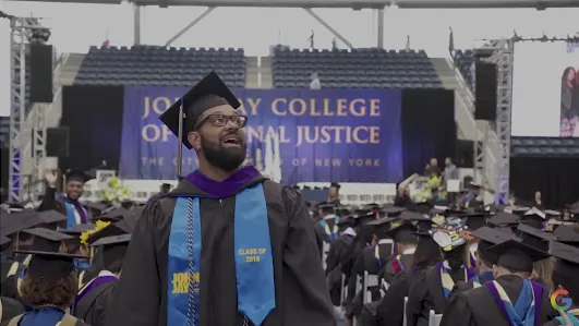 A joyful graduate from the John Jay College Class of 2019 looks upward with a smile during a commencement ceremony. He is wearing a black graduation gown, a mortarboard cap, and a light blue stole that reads "Class of 2019." In the background, a large blue banner for the John Jay College of Criminal Justice and rows of seated graduates are visible.