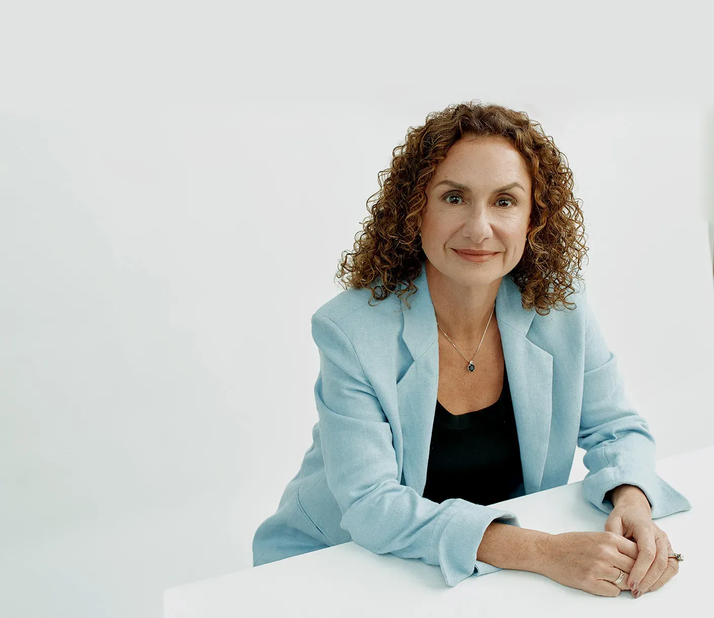 A professional portrait of Lila Ibrahim, Chief Operating Officer of Google DeepMind. She has curly brown hair and is wearing a light blue blazer over a black top, sitting with her hands clasped on a white surface against a clean white background.