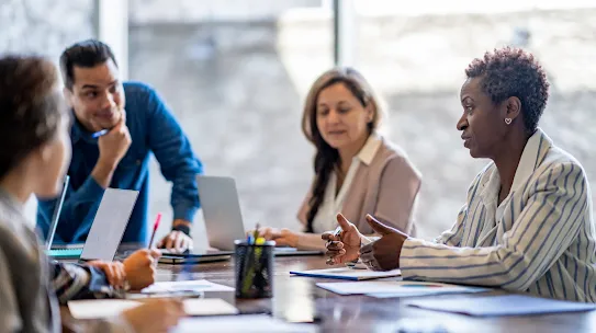A woman speaking in a professional meeting while seated at a conference table with colleagues.