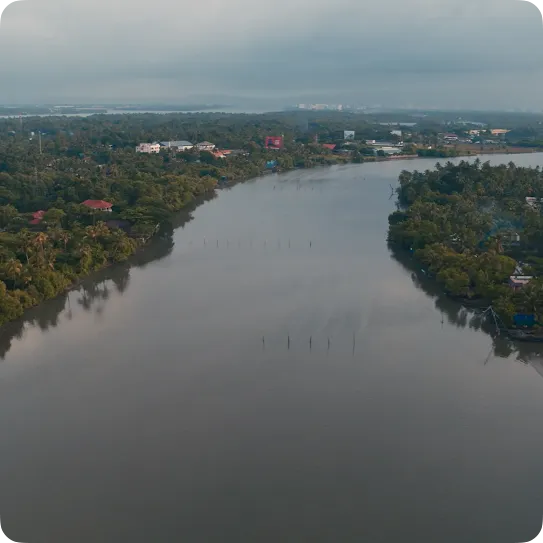High-angle aerial view of a wide, calm river winding through a dense tropical landscape filled with green trees and small buildings under an overcast, cloudy sky.