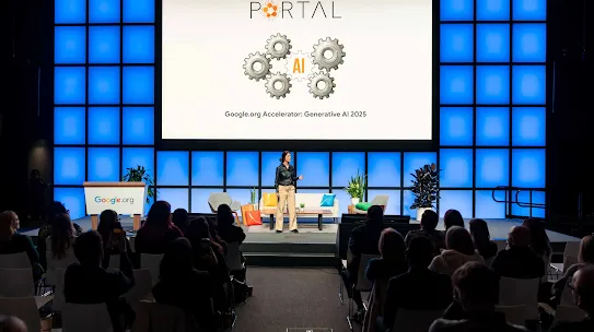 A wide shot of a speaker on a stage in front of a seated audience. The backdrop features a grid of glowing blue squares and a large central screen displaying the word "PORTAL" above an illustration of gears with "AI" in the center. The text on the screen reads "Google.org Accelerator: Generative AI 2025." To the left of the speaker is a podium with the Google.org logo.