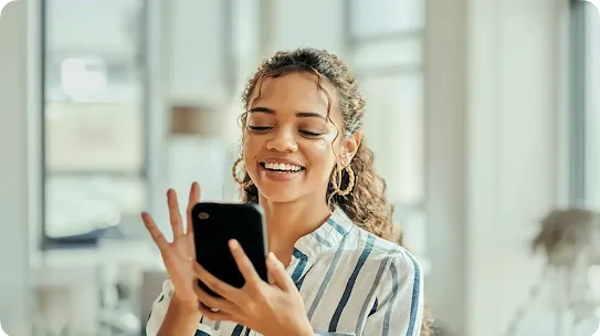 A smiling young woman with curly hair and gold hoop earrings holding a smartphone, waving at the screen during a video call.