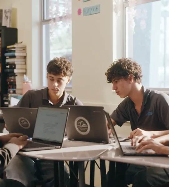 Four high school students working on laptops at a group of desks in a sunlit classroom, focusing on digital learning.