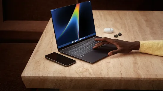 A Googlebook laptop open with a phone and earbuds nearby.