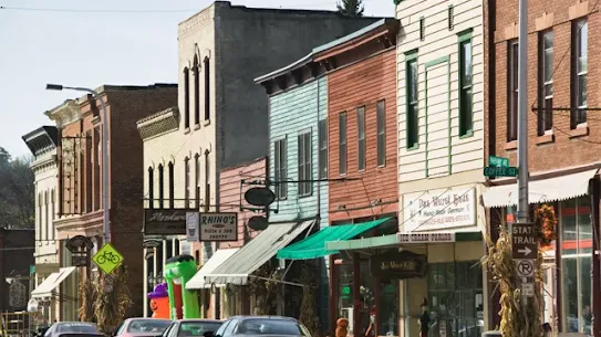 A row of colorful historic buildings along a sunny American Main Street. Storefronts include local businesses like a pizza shop and an ice cream parlor, with autumn decorations like pumpkins and corn stalks on the sidewalk.