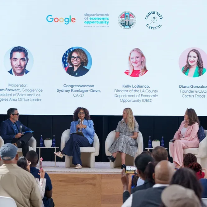 A panel of four individuals sit on a stage, addressing an audience with a screen that displays Google, Los Angeles County Dept of Economic Opportunity