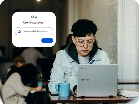 A person wearing glasses and a denim jacket looks at a Samsung Chromebook in a cafe. A digital overlay shows a Google "Use this passkey?" sign-in prompt for elisa.g.beckett@gmail.com.