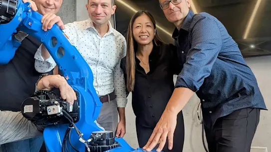 A group portrait of four people from the robotics software company Intrinsic posing with a large, bright blue industrial robotic arm in an office setting. They are smiling and touching the base of the machine, celebrating the news of joining Google to further develop industrial robotics software.