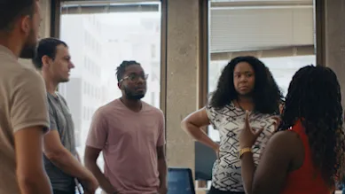 five people standing in a circle talking, two backs are to the camera